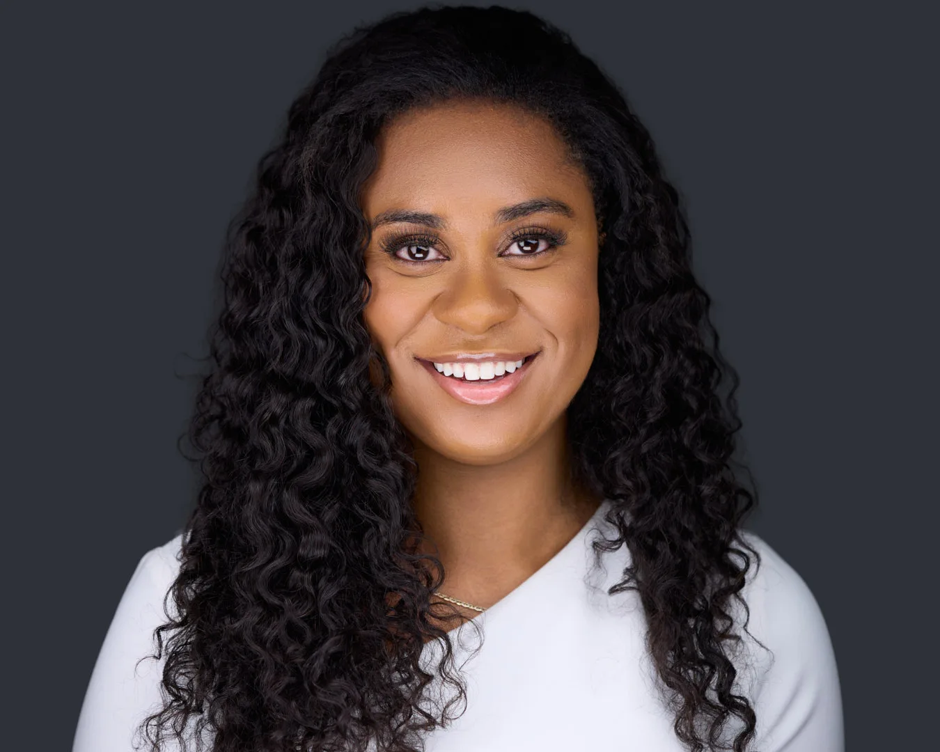 Professional studio headshot of a smiling woman with long curly hair in a white top against a charcoal background, the kind of current, well-lit image that performs on LinkedIn