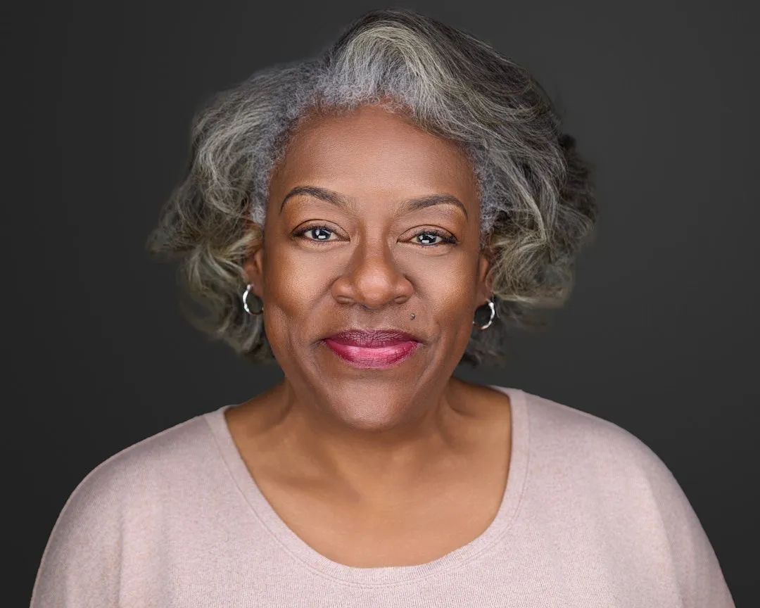 Close-up portrait of an African-American woman with gray curly hair, wearing a beige top, earrings, and lipstick, against a dark background.