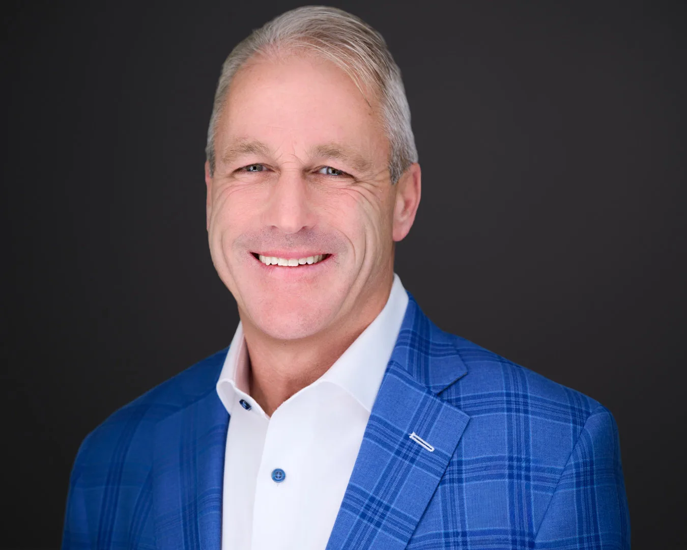 Professional headshot of a silver-haired executive in a blue plaid blazer on a dark studio backdrop
