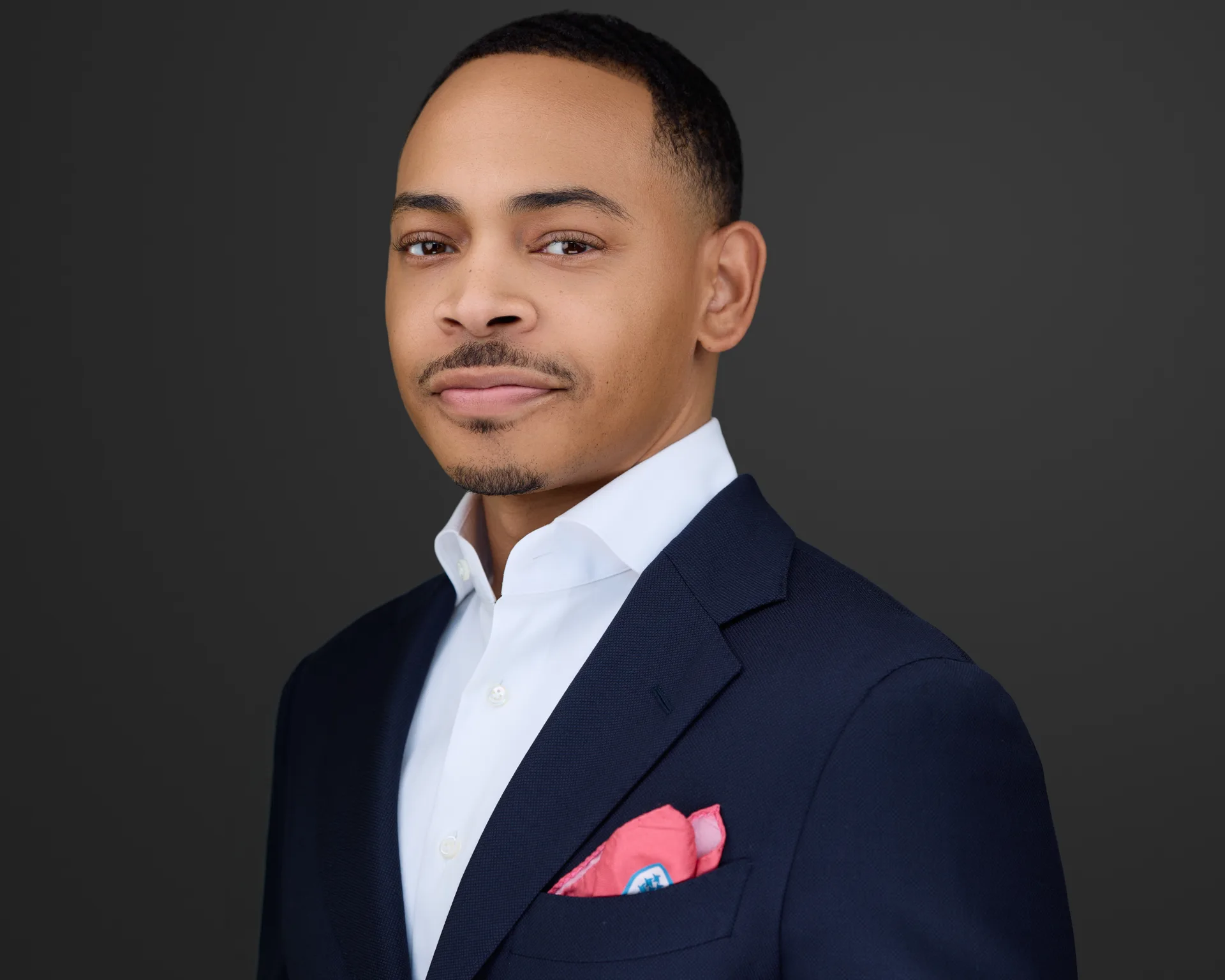 Professional headshot of a businessman in a navy blazer with a pink pocket square on a dark studio backdrop