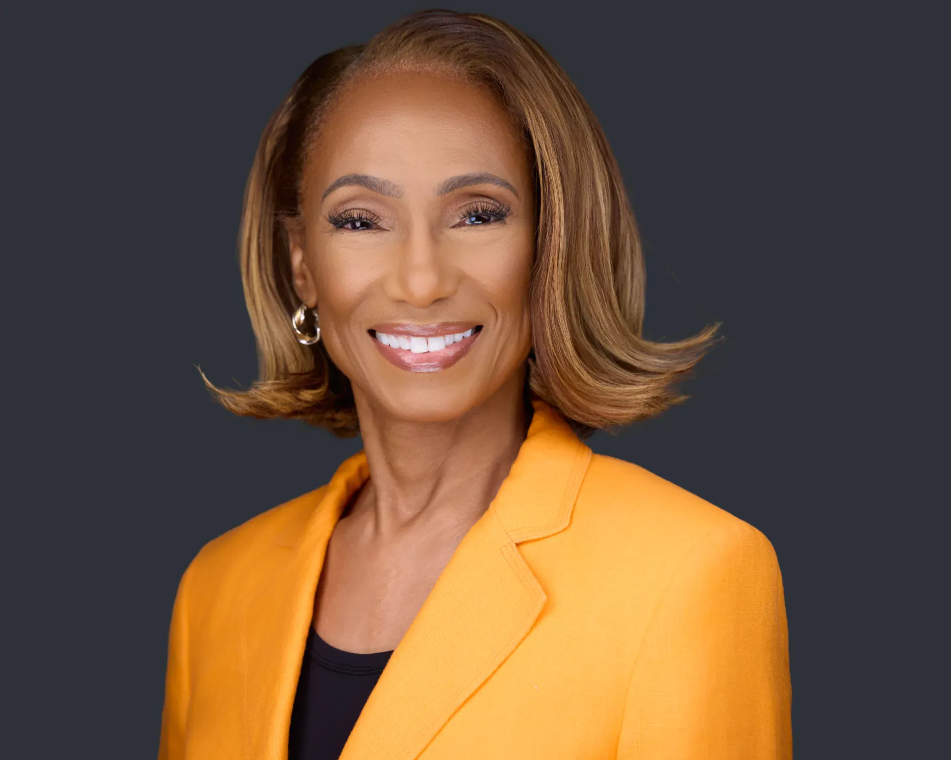 Professional headshot of a smiling executive in a marigold blazer and gold hoop earrings on a dark studio backdrop