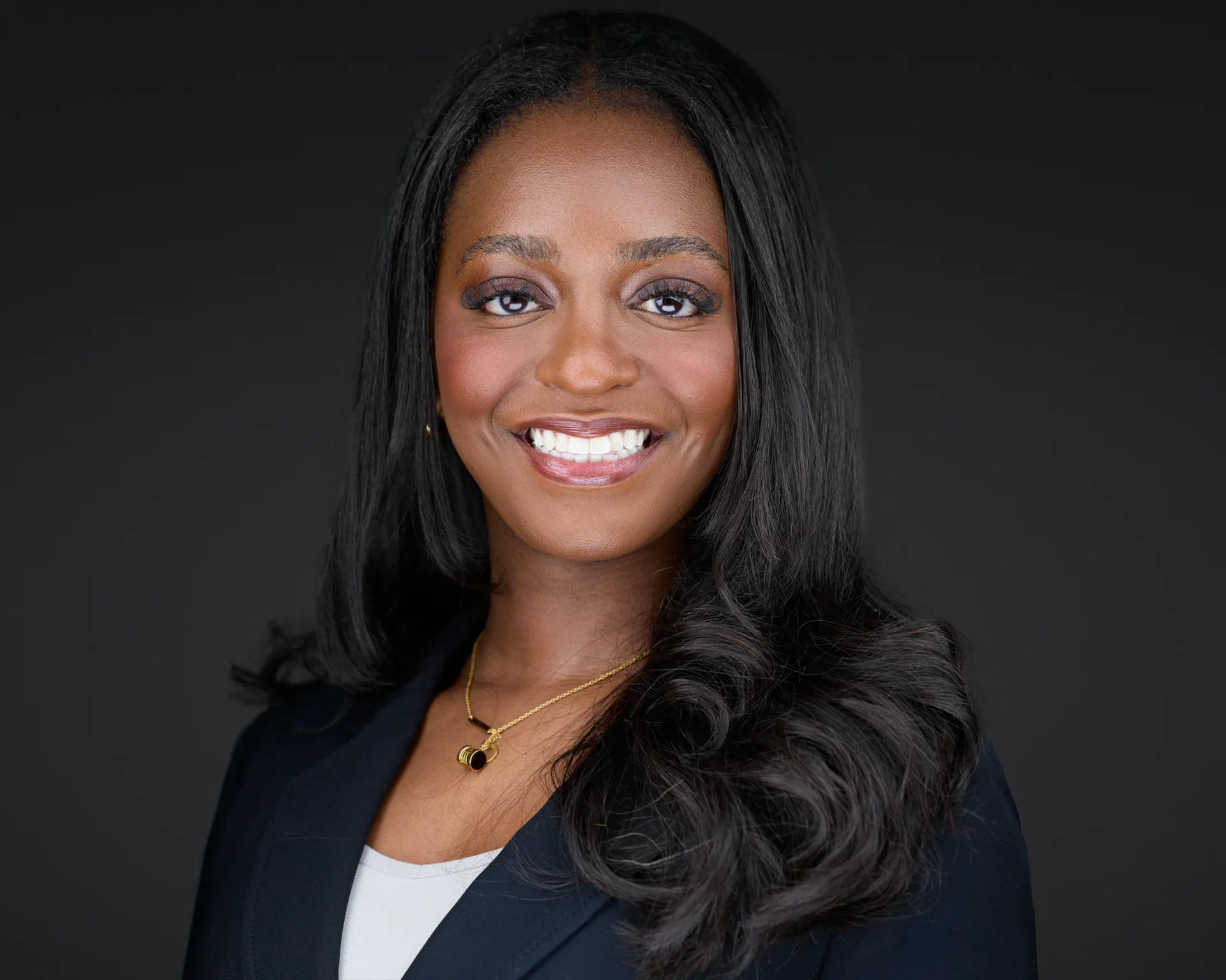 Professional headshot of a businesswoman in a dark blazer and gold necklace on a dark studio backdrop