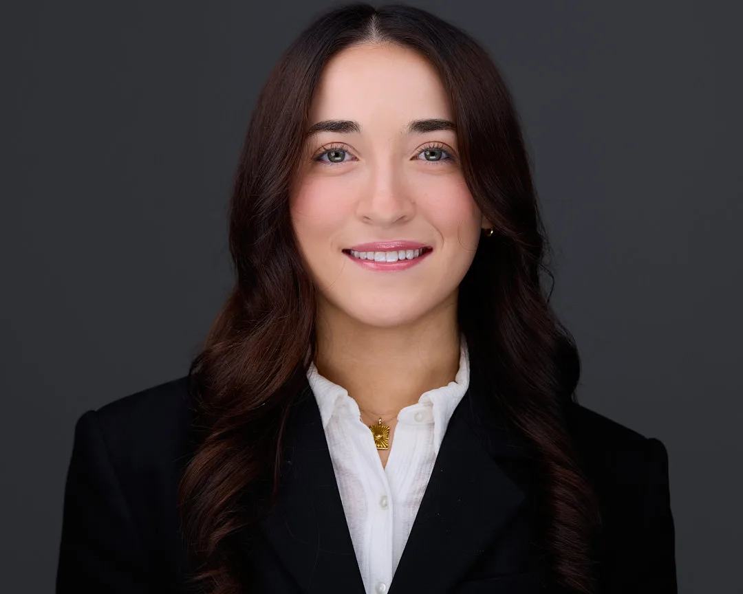 Professional headshot of a businesswoman in a black blazer and white shirt with a gold necklace on a dark studio backdrop